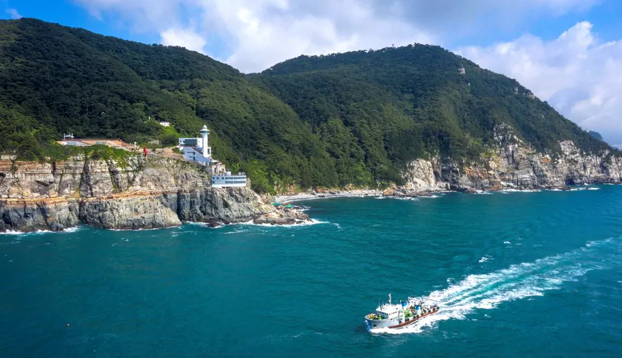 釜山・太宗台の海食崖と影島灯台、森、沖を進む遊覧船